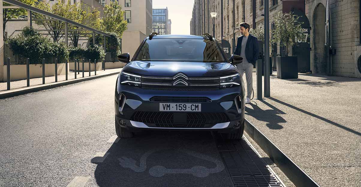 Citroen_C5_Aircross_Hybrid_Rechargeable_urban Brünetter Mann in Business-Look lädt C5 Aircross Hybrid 2022 an Straße in urabnem Umfeld.